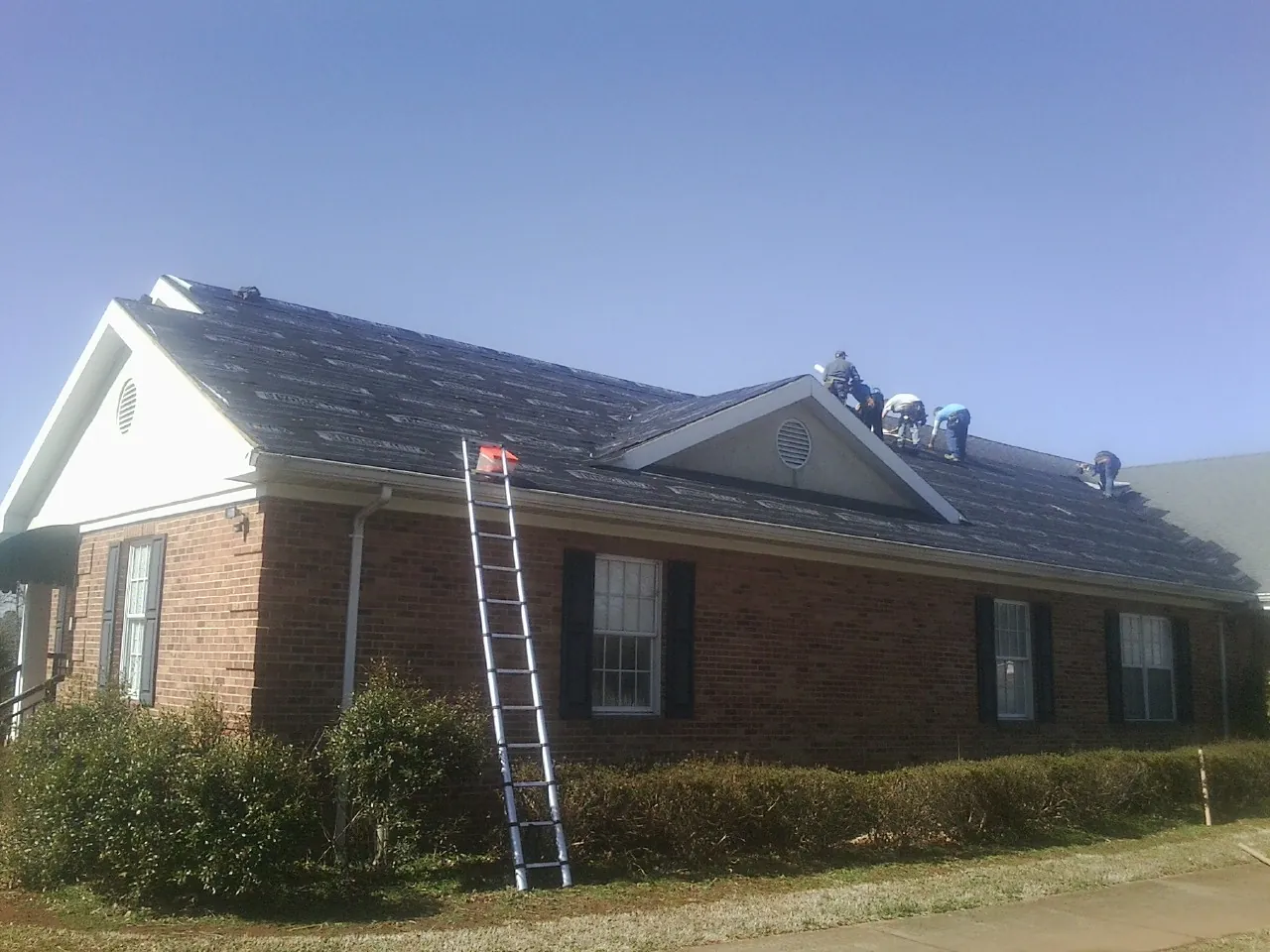 Crew tearing shingles off a brick ranch roof with a ladder set — Emergency Roof Repair work by our crew in Altus