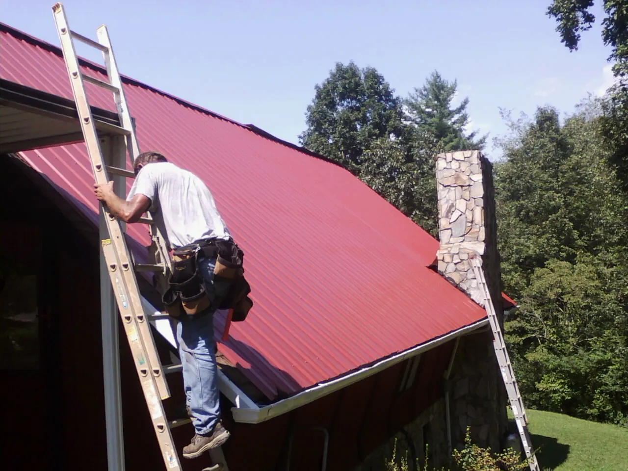 Roofer on a ladder beside a red metal roof and stone chimney — Soffit & Fascia Repair by Roofing Brothers in Altus